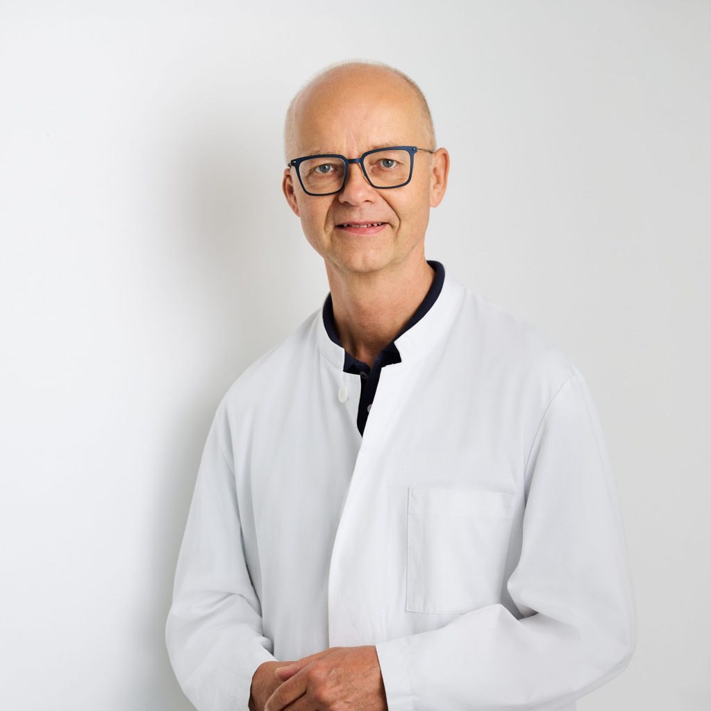 Sie sehen ein Portraitfoto eines Mannes mittleren Alters. Er trägt einen weißen Arztkittel und lächelt in die Kamera. Seine Haare sind weiß, kurz und dünn. Er trägt eine eckige Brille mit dunklem Gestell.