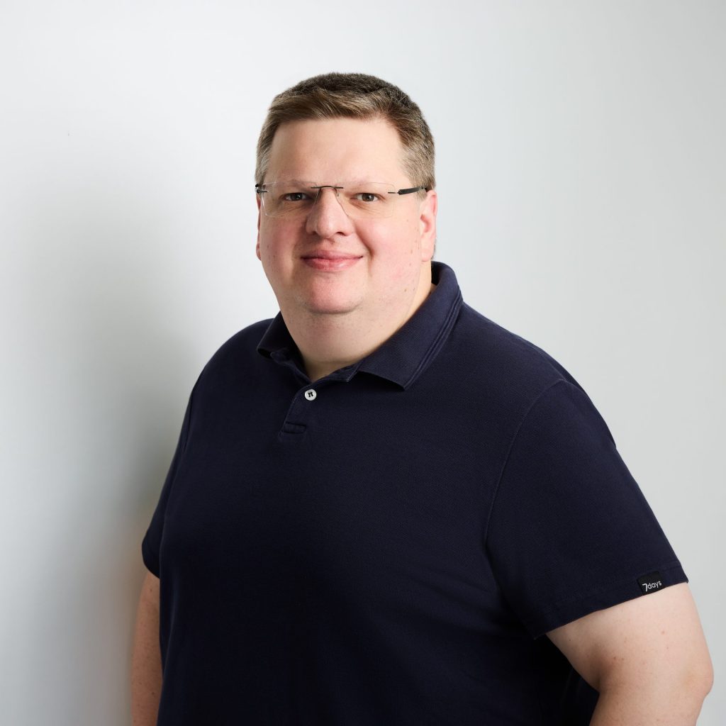 Sie sehen einen kräftigen Mann mittleren Alters in die Kamera lächeln. Er trägt kuzes blondes Haar, eine Brille und ein dunkelblaues Poloshirt.