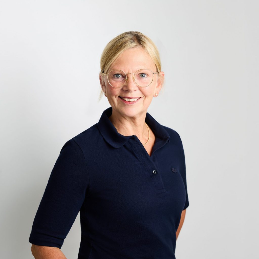Sie sehen das Portraitfoto einer lächelnden Frau mittleren Alters mit blonden, zum Pferdeschwanz zusammengebundenen Haaren. Sie trägt eine große runde Brille mit goldenem Rahmen und ein dunkelblaues Poloshirt.