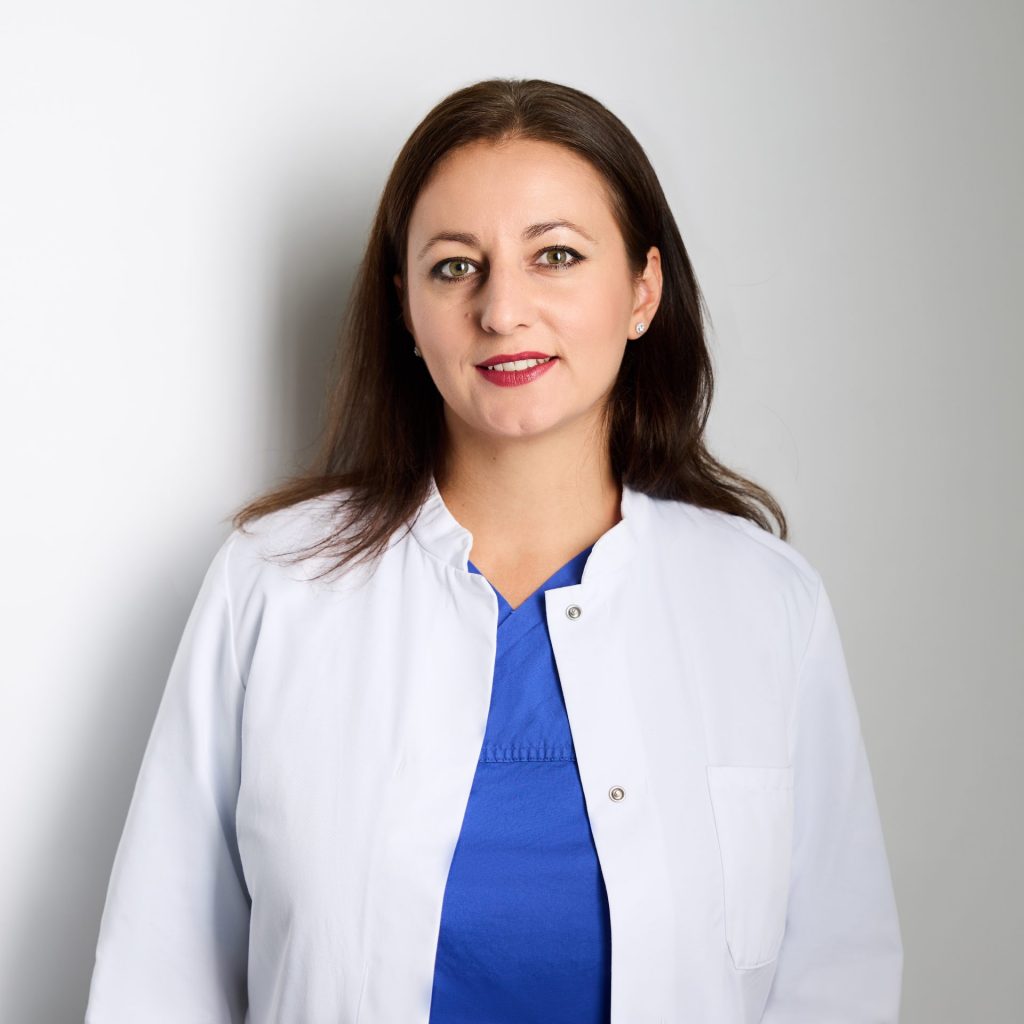Sie sehen eine lächelnde junge Frau in einem blauen Top und weißen Arztkittel. Das braune Haar ist offen und schulterlang.