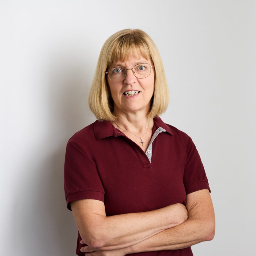 Sie sehen eine Frau mittleren Alters in bordeauxrotem Poloshirt. Die blonden Haare mit Pony sind schulterlang. Sie trägt eine große ovale Brille mit hellem Gestell.