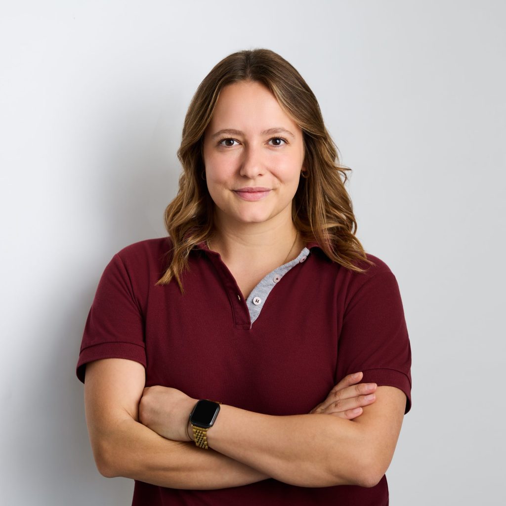 Sie sehen eine lächelnde junge Frau in bordeauxrotem Poloshirt und braunen lockigen Haaren, die etwas länger als bis zur Schulter gehen.