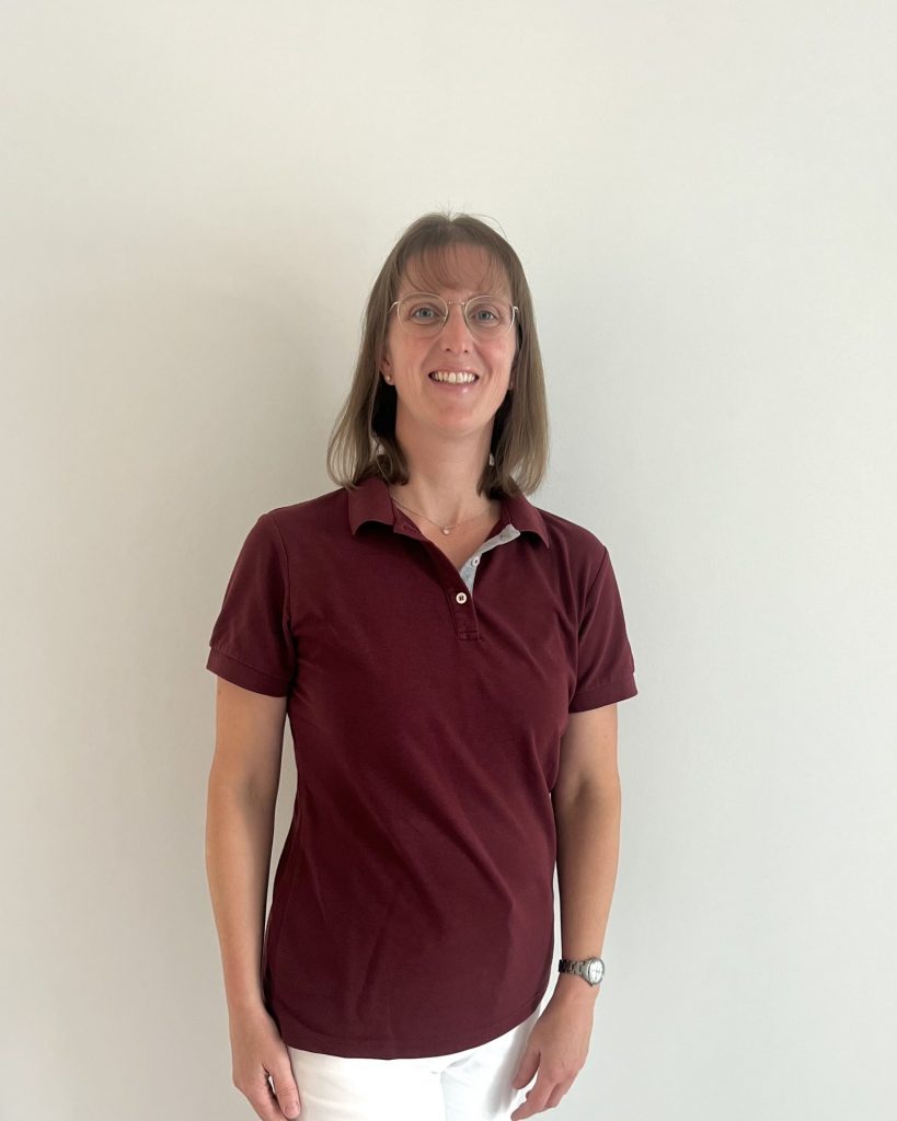 Sie sehen eine lachende Frau mittleren Alters in bordeauxrotem Poloshirt und weißer Hose. Die braunen Haare mit Pony sind dünn und schulterlang. Sie trägt eine ovale Brille mit silbernem Gestell.