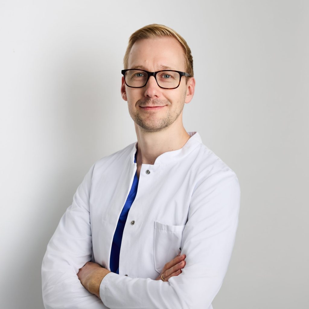 Sie sehen das Portraitfoto eines jungen Mannes in weißem Arztkittel der freundlich lächelt. Das dunkelblonde Haar ist kurz, er trägt einen kurzen Bart und Brille.