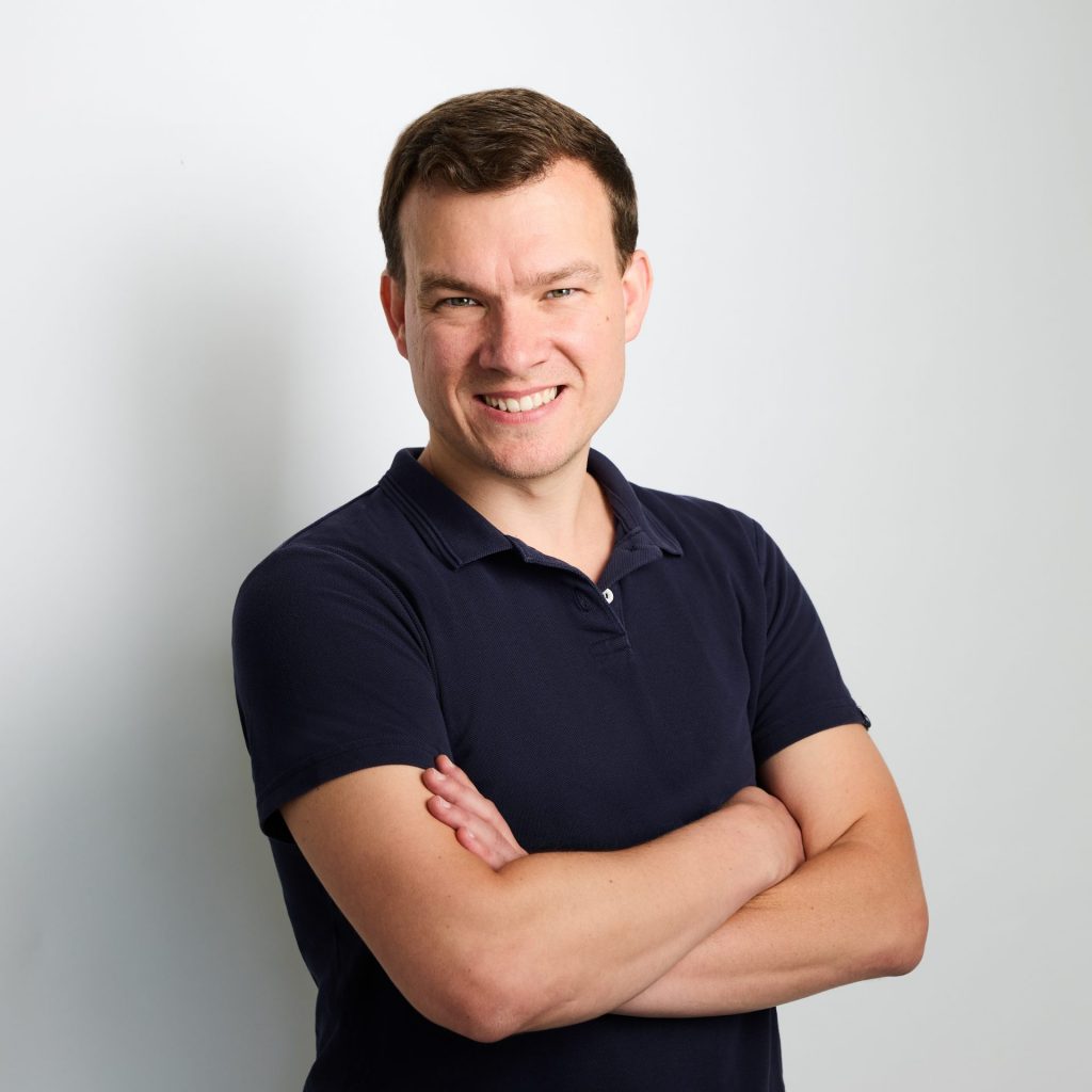 Sie sehen das Portraitfoto eines jungen Mannes in dunkelblauem Poloshirt, der freundlich lächelt. Das dunkelblonde Haar ist kurz, die Arme verschränkt.